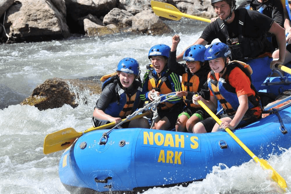 A group of kids and an adult enthusiastically navigate a river in a blue raft labeled "Noah's Ark."