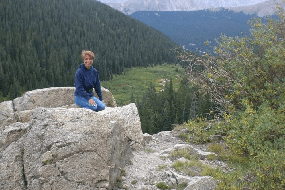 A woman sits on a large rock in a mountainous landscape surrounded by evergreen trees.