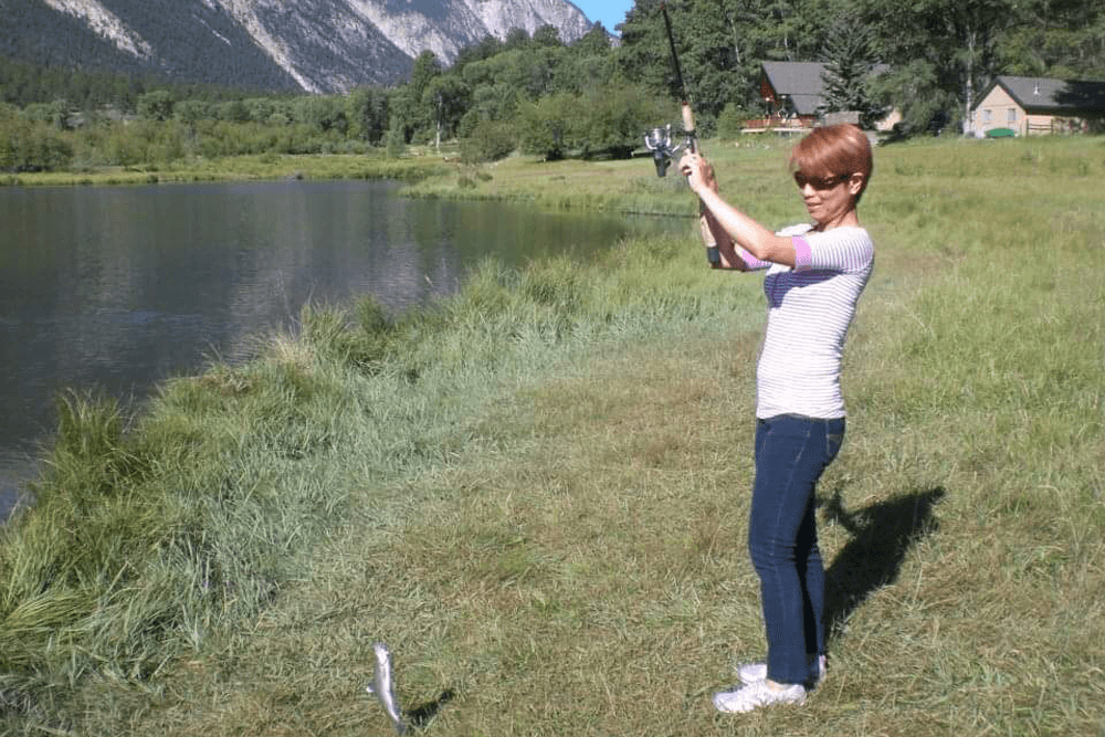 A woman stands by a lake, holding a fishing pole as a fish jumps out of the water.