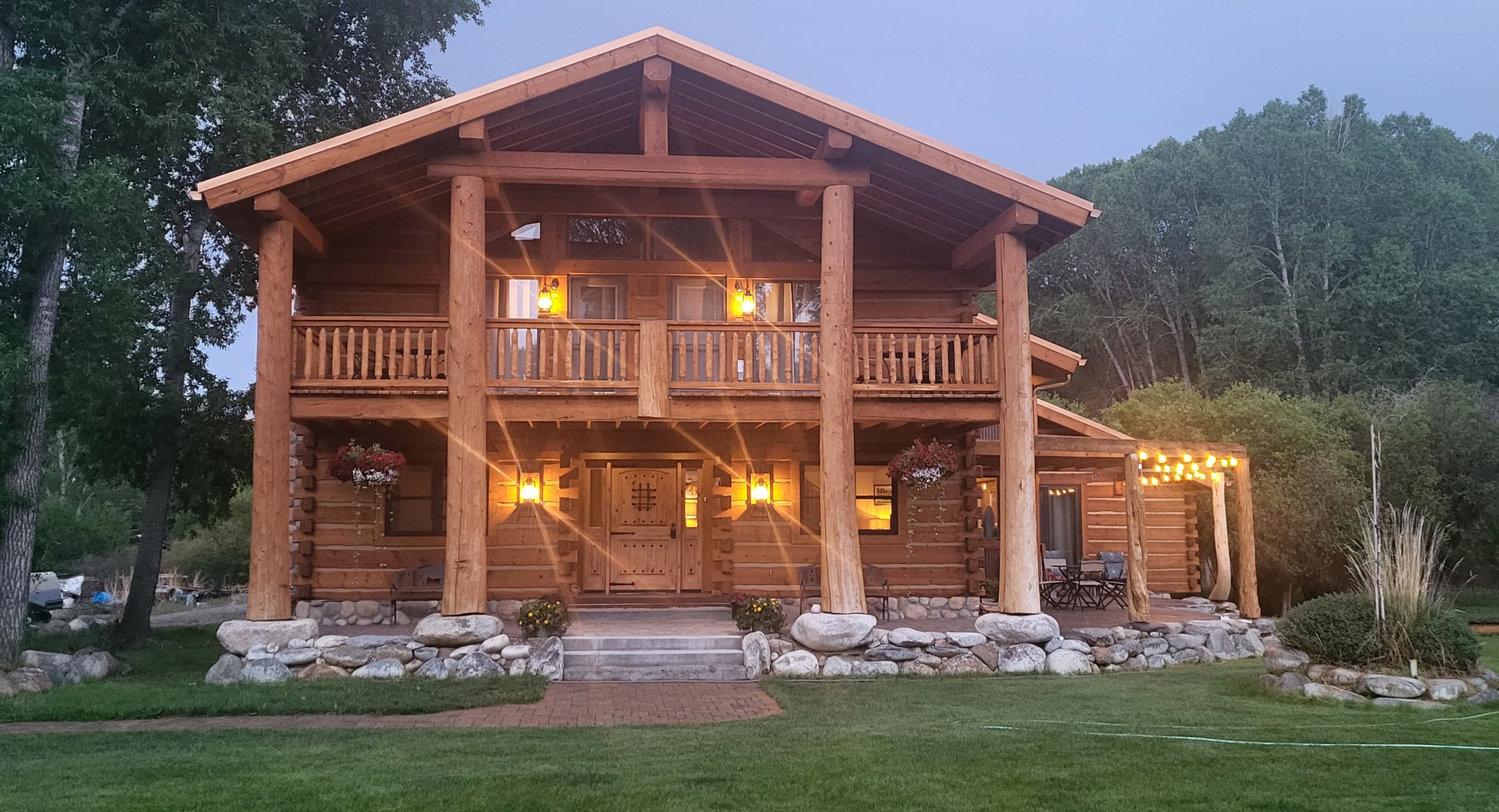 A log cabin house with a spacious porch, surrounded by trees and a grassy lawn, illuminated in the evening.